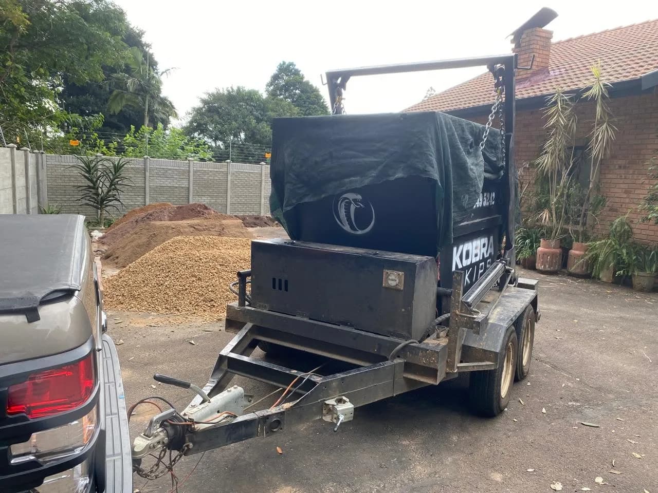 Kobra Skip covered and ready for collection from a residential driveway, mulch pile visible from a garden project.