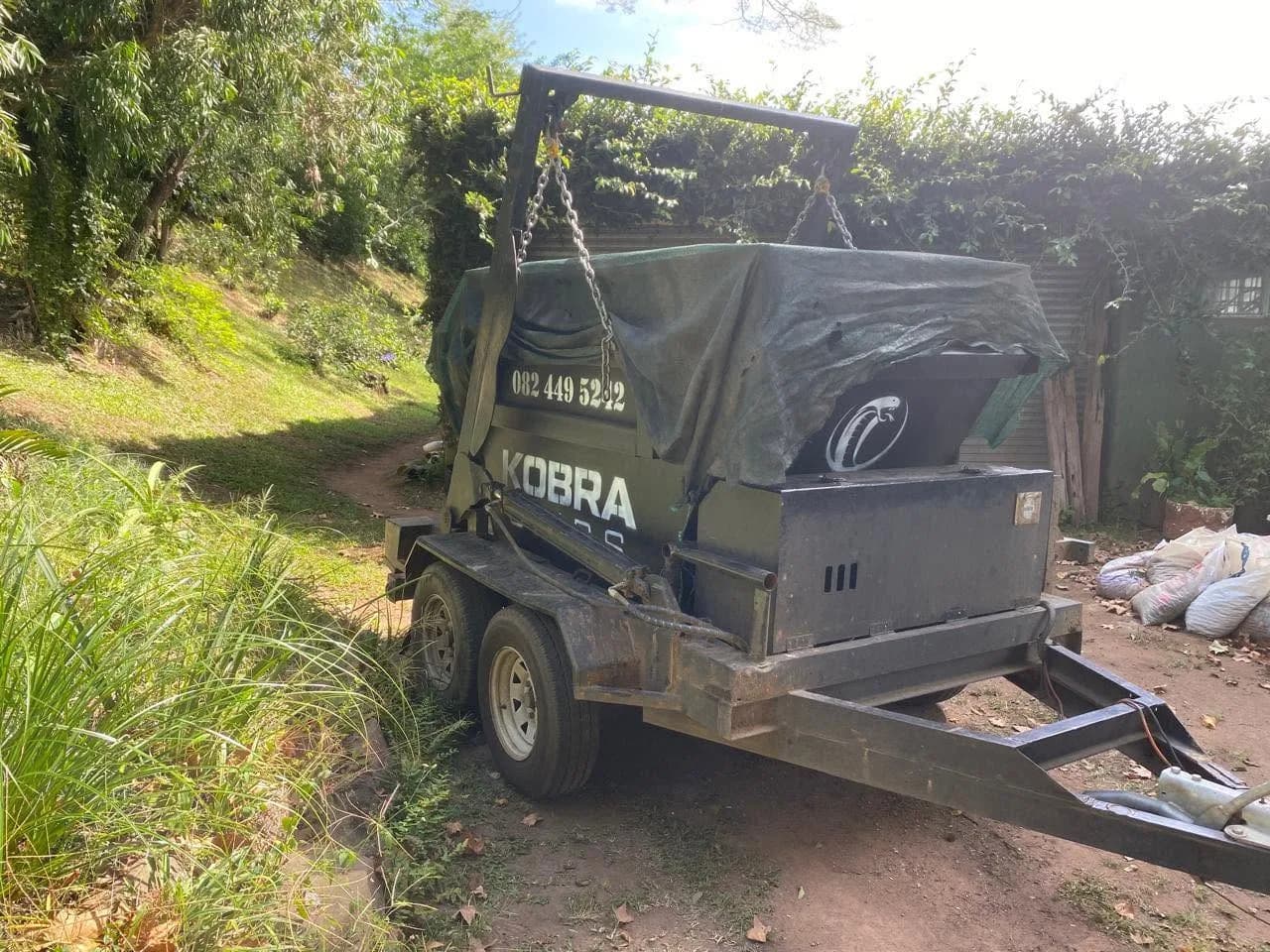 Kobra Skip on trailer parked on a dirt track, surrounded by dense green bush after a garden clearance.